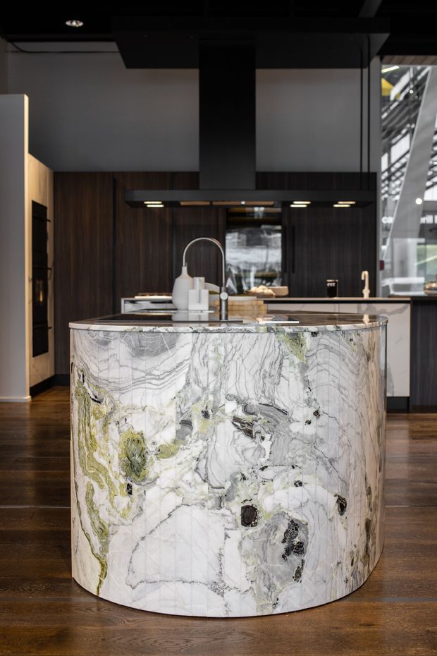 A dramatic contemporary kitchen centered on a large curved island clad in richly veined natural stone with white, grey, black, and subtle green tones. The island includes an integrated sink and brushed metal tap, with dark timber cabinetry, a dark ceiling, black rangehood, and warm timber flooring. A second pale benchtop and cabinetry run behind the island, with glazing visible to the right.