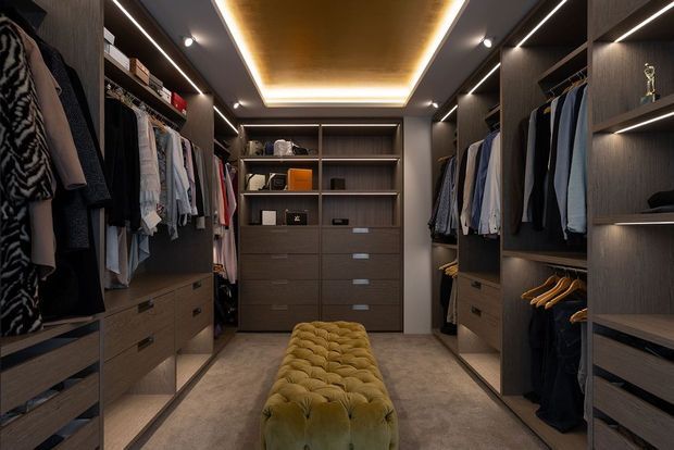 A luxurious walk-in wardrobe with dark wood built-in cabinetry, open hanging rails, drawers, shelving, integrated strip lighting, and a central tufted upholstered bench. The room has a warm, moody atmosphere with a carpeted floor and a recessed illuminated ceiling feature.