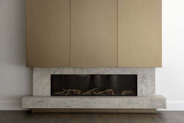 Minimalist contemporary fireplace feature wall with a wide linear firebox set into a pale stone surround and hearth. Large matte taupe-brown built-in cabinetry spans above the fireplace, with neutral plaster walls and dark timber flooring.