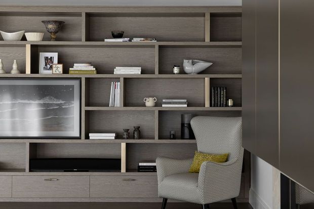 A refined built-in media and display wall in a dark grey-brown wood finish, with open shelving, lower cabinetry, books, ceramics, framed photographs and a television. A patterned upholstered armchair with a mustard cushion sits in front, while a dark fireplace or architectural element is partially visible at right. The composition is tailored, layered and contemporary with warm neutral tones.
