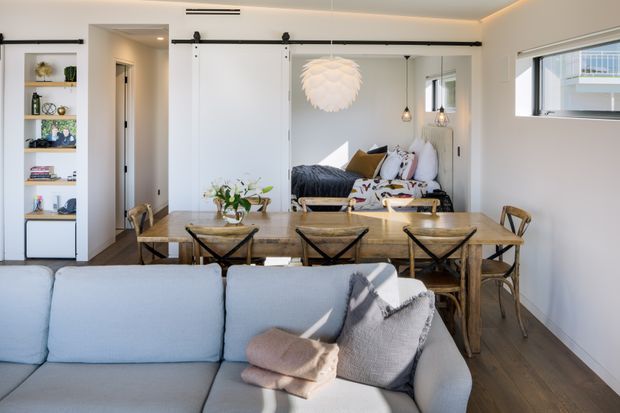 Bright open-plan living and dining interior with a pale blue sectional sofa in the foreground, a rustic timber dining table and cross-back chairs, and a compact lounge area beyond. White walls, timber flooring, black barn-style sliding doors and shelving, layered cushions, and a sculptural pendant create a relaxed contemporary coastal character.