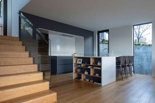 Contemporary open-plan kitchen and living area with a floating timber staircase, glass balustrade, dark built-in cabinetry, white island bench, timber shelving, and a compact dining or breakfast bar. Large windows provide abundant natural light, while timber flooring and cabinetry add warmth to the restrained palette.