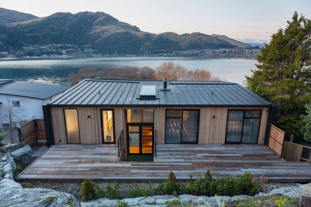 Exterior view of a compact single-storey contemporary home with vertical timber cladding, a dark standing-seam metal gable roof, large glazed openings, and a broad timber deck. The house is set on a rocky elevated site overlooking a lake and surrounding mountainous terrain.