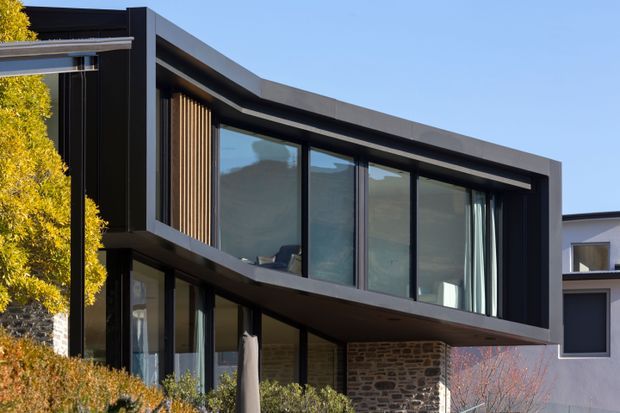 Contemporary two-storey residential facade with a bold dark framed upper-level glazed volume projecting over a stone-clad lower section. Floor-to-ceiling windows, vertical timber screening, deep roof eaves, and strong horizontal and rectilinear forms define the exterior. A neighbouring light-coloured building and landscaped planting are visible around the house.