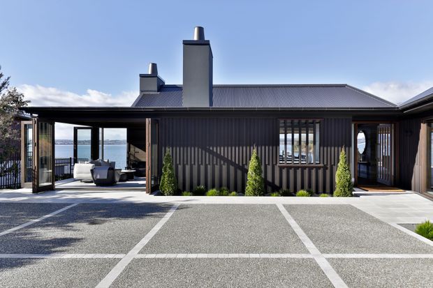 Exterior view of a contemporary single-storey home with dark vertical ribbed cladding, a low pitched metal roof, prominent chimney structures, and extensive glazed openings. A covered outdoor seating area and waterfront view are visible through the left opening, with a paved courtyard or driveway in the foreground and structured planting along the facade.