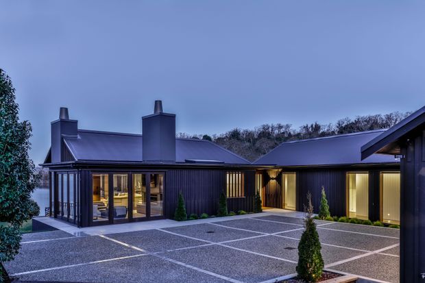 A dusk exterior view of a contemporary single-storey residence arranged around a broad paved courtyard. The dark vertical-clad building has low pitched metal roofs, prominent chimney forms, extensive glazed living areas, warm interior lighting, and carefully planted conical shrubs. The courtyard features dark gravel or textured paving divided by light linear bands.