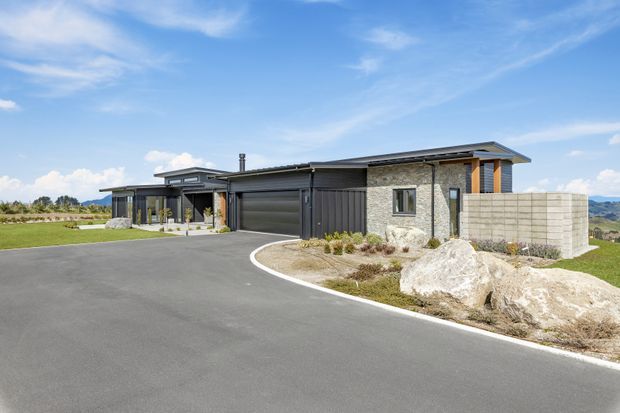Single-storey contemporary residential home viewed from the driveway, with a low-pitched roof, dark vertical cladding, natural stone wall sections, concrete block screening, broad garage doors, and sheltered outdoor entry areas. The landscaped frontage includes large rocks and sparse planting, with open rural or semi-rural surroundings visible beyond the property.