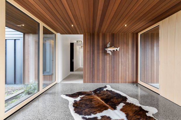 A refined residential entry hall with a warm timber-lined ceiling and feature wall, broad glazed openings to a small courtyard, pale built-in cabinetry, polished grey terrazzo flooring, and a cowhide rug. Recessed spotlights and a sculptural wall-mounted shelf add understated detail, while a corridor extends toward the rear of the home.