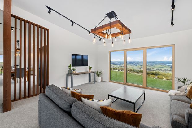 Bright contemporary living room with a large grey sectional sofa, low black-framed coffee table, wall-mounted television, and a slim console. Full-height timber-framed glazing opens views toward rolling green countryside. A timber slatted room divider and exposed timber post define the entry edge, while a timber-and-black-metal pendant with multiple hanging bulbs and black track lighting provide illumination. The room has light carpet, white walls and ceiling, and a calm neutral palette.