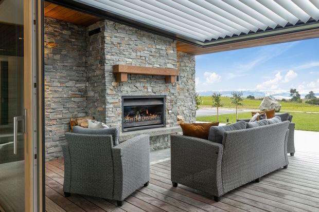 A covered outdoor living area with a stone-clad fireplace, timber ceiling and decking, a louvred roof, and woven outdoor seating arranged around the fire. The space opens onto a broad rural landscape and is brightly lit by daylight.