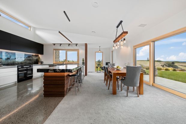 A bright, open-plan kitchen and dining area with polished concrete flooring, white and black cabinetry, a timber kitchen island, stone benchtops, and a timber dining table. Large windows and glazed doors provide expansive views of a coastal landscape. Linear pendant lighting and exposed timber accents add contemporary character.