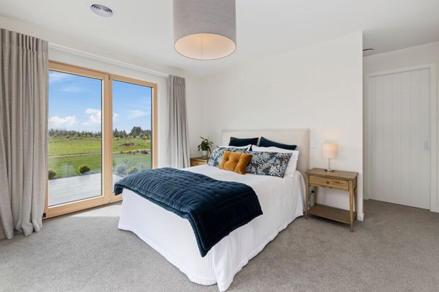 A bright, inviting bedroom with a white upholstered bed, white bedding, navy quilt, and patterned decorative cushions. Large timber-framed glazed doors open to a scenic green landscape, while full-height neutral curtains, pale carpet, a simple timber bedside table, and a soft pendant light create a calm contemporary interior.