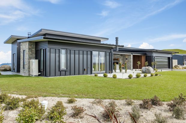Single-storey contemporary residential home viewed from the front, with a layered low-pitched roofline, dark vertical metal-clad facade, grey masonry and stone feature walls, extensive glazing, and a broad lawn with simple landscaped planting. The building has a clean horizontal composition and sheltered entry or outdoor living areas.