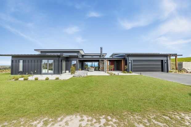Wide exterior view of a contemporary single-storey residence with a low-slung, multi-wing flat roof, dark vertical cladding, extensive glazing, a central recessed entry or courtyard, and an attached double garage. The home sits behind a broad manicured lawn with a paved driveway and sparse landscaping under a bright blue sky.