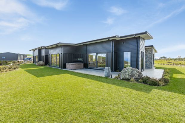 Single-storey modern house viewed from the landscaped front lawn, with dark horizontal exterior cladding, a low-pitched roof, extensive floor-to-ceiling glazing, and a paved outdoor entertaining area. Natural stone accents and neatly planted garden beds surround the contemporary dwelling.