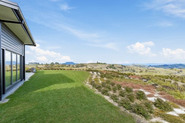 A contemporary single-storey house is shown from the side, with horizontal weatherboard cladding, extensive floor-to-ceiling glazing, and a projecting roof edge. The building opens onto a neatly mown lawn and a naturalistic coastal or rural landscape with low scrub, rolling fields, and distant hills under a bright blue sky.