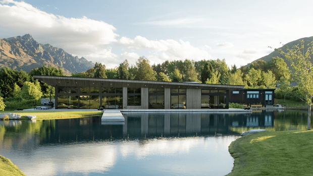 A long, low modern pavilion-style residence set beside a large reflecting or swimming pool, with extensive floor-to-ceiling glazing, slender dark structural framing, and a flat roof. A mountainous landscape and mature trees form the background, while the building’s interior is partially visible through the glazing.