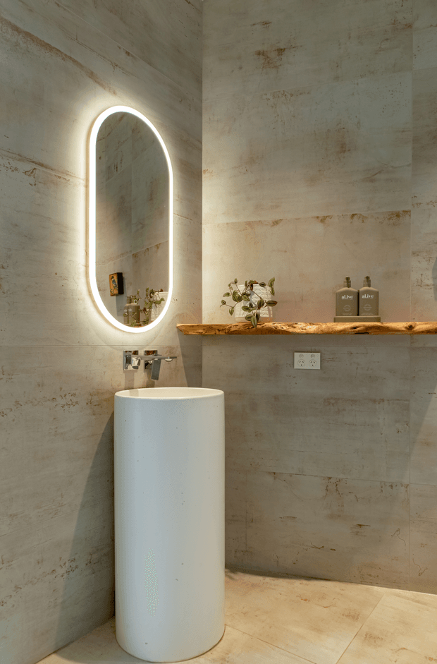 A compact, refined bathroom featuring large warm grey-beige stone-look wall and floor surfaces, a freestanding cylindrical white basin with wall-mounted chrome tapware, and a tall arched backlit mirror. A rustic natural-edge timber shelf spans the wall, holding small plants and matching amenity bottles. The space combines minimalist fixtures with natural, weathered textures and warm ambient lighting.