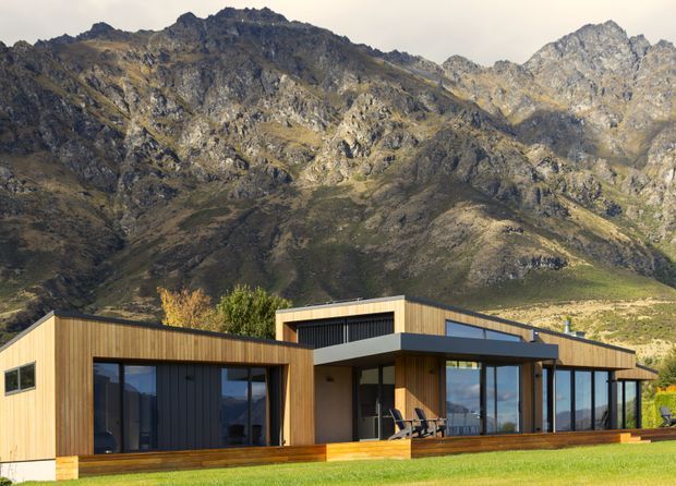 A low-slung contemporary timber-clad house set against a dramatic rugged alpine mountain landscape. The home features broad floor-to-ceiling glazing, dark vertical wall panels, projecting flat roof forms, and a timber deck opening onto a lawn.