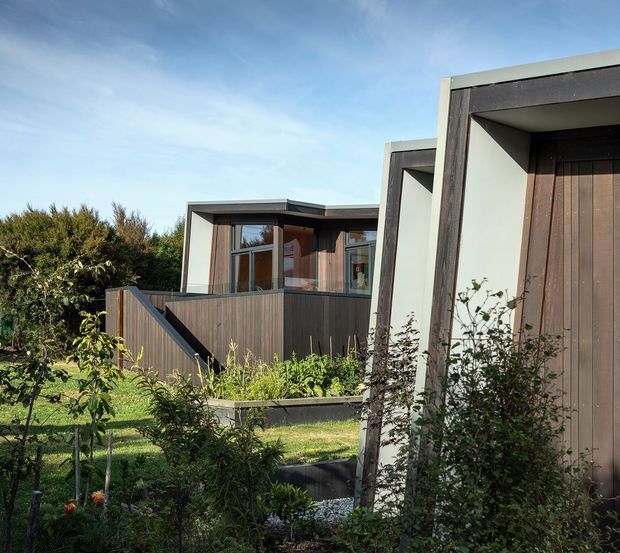 Exterior view of a contemporary low-rise residential building with layered rectangular volumes, dark timber cladding, pale rendered or concrete-framed openings, and extensive glazing. A raised timber deck and stair connect the building to a landscaped garden with shrubs and young trees. The composition emphasizes modern geometric forms and a natural outdoor setting under a clear sky.