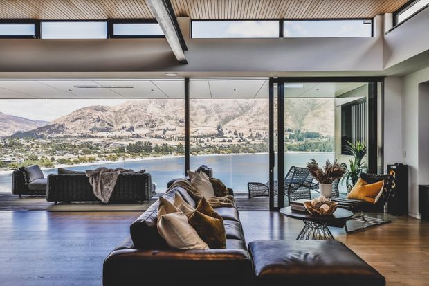 A spacious contemporary living room with expansive floor-to-ceiling glazing and open glass doors overlooking a lake and mountainous landscape. The room features dark leather sofas and ottoman seating, woven lounge chairs, a low coffee table, indoor plants, and a warm timber-lined ceiling with exposed structural beams. The design creates a strong indoor–outdoor connection and a relaxed, view-focused atmosphere.