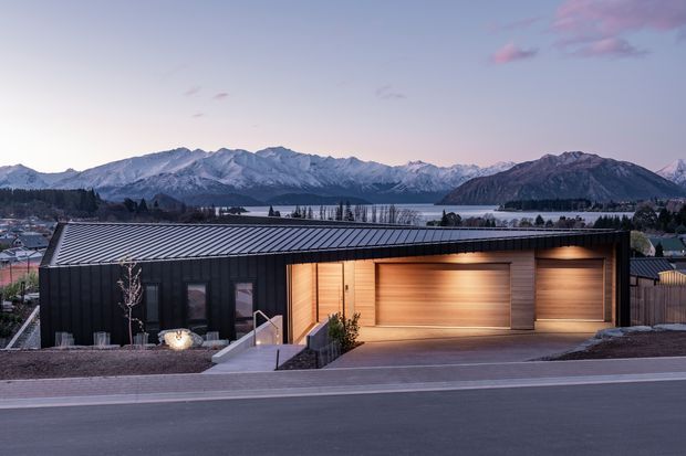 Contemporary single-storey residence viewed from across the street at dusk, with a dark metal-clad facade, low sloping roof, and warm timber-lined recessed garage and entry volumes. The home sits in a developed lakeside or mountain-town setting, with snow-capped mountains and a body of water visible in the background. A paved street and landscaped frontage frame the foreground.