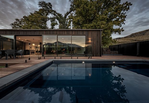 A single-storey modern residence viewed from the pool, with a dark timber-clad facade, extensive floor-to-ceiling glazing, and a covered outdoor entertaining area. The reflective swimming pool occupies the foreground, while a paved pool deck, black fencing, mature trees, and a dramatic hillside setting frame the home under an overcast sky.
