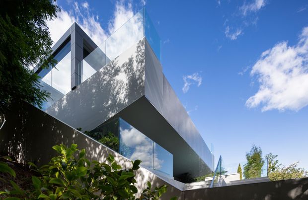 Exterior architectural photograph of a highly contemporary, angular building with a dramatic cantilevered upper volume, extensive reflective glazing, smooth light-grey cladding, and sharp geometric forms. The structure is viewed from a landscaped urban or suburban setting beneath a bright blue sky, with trees and planting partially screening the lower facade.
