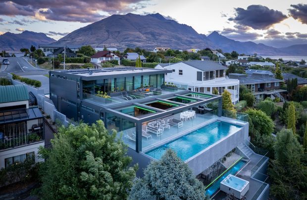 Aerial view of a contemporary multi-level residence set within a suburban landscape, with a large rooftop terrace, outdoor dining area, and elevated swimming pool. The home features strong geometric forms, extensive glazing, dark cladding, flat roofs, and landscaped surroundings. Mountains and a body of water are visible in the background.
