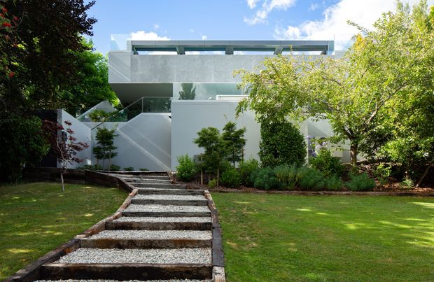 Exterior view of a contemporary multi-level residence set within a landscaped garden. A stepped gravel-and-timber walkway leads toward angular white rendered walls and a projecting upper volume with broad grey concrete-like surfaces and horizontal glazing. Mature trees and lawns frame the geometric facade.