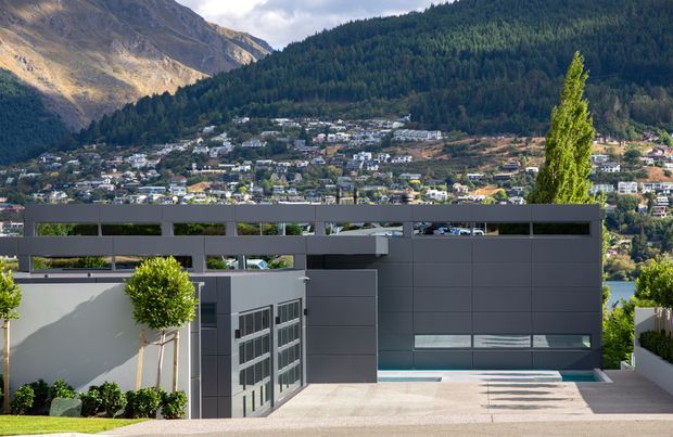 A contemporary multi-level residential exterior on a sloped site, featuring dark grey horizontal cladding, broad bands of glazing, a flat roofline, and a paved driveway or terrace in the foreground. The property overlooks a dense hillside settlement and dramatic forested mountain landscape, with clipped ornamental trees and planted borders around the building.