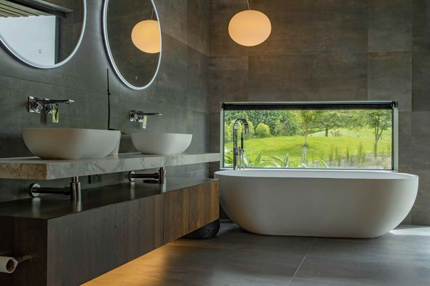 Contemporary bathroom with a freestanding oval bathtub positioned beside a large horizontal window overlooking greenery. A floating dark timber double vanity with two white vessel basins sits against large dark grey concrete-look wall panels, complemented by round mirrors with illuminated edges and warm globe pendant lights. The room has a refined, moody yet naturally connected atmosphere.
