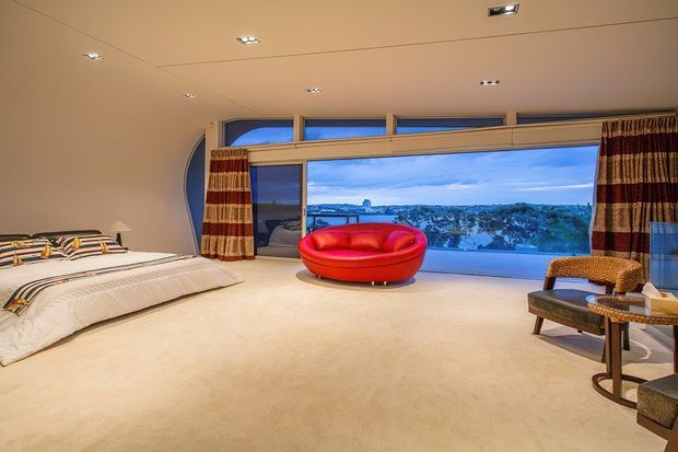Spacious bedroom with a low-profile bed, cream carpet, striped floor-to-ceiling curtains, and a vivid red circular lounge chair. A wide picture window overlooks a leafy landscape and distant water or cityscape, while recessed ceiling lights and a curved blue-glass architectural detail add a distinctive modern character.