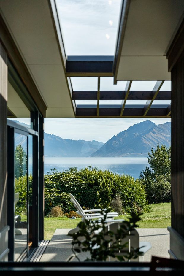 A view from inside a contemporary glazed living space toward a landscaped lawn and a large alpine lake framed by rugged mountains. Floor-to-ceiling dark-framed glazing and a partially covered outdoor terrace create a strong indoor-outdoor connection, with a skylit or glazed canopy overhead and lounge chairs outside.