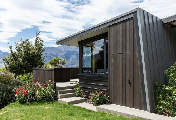 A modern single-storey home exterior set in a landscaped garden, with a low-slung projecting roof, dark vertical timber cladding, corrugated dark metal wall panels, extensive glazing, and concrete steps leading to the entry. Mountain scenery is visible in the background.