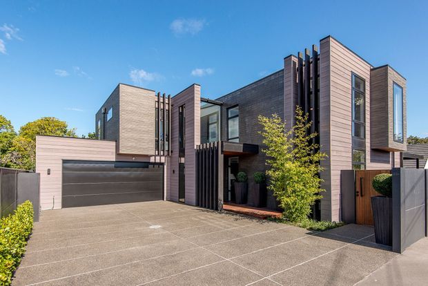 Contemporary multi-level residential facade with a broad concrete-paved driveway, integrated double garage, projecting box forms, vertical dark slat screens, and extensive horizontal timber-look cladding in muted grey and mauve tones. Large windows and a recessed entry are framed by minimalist landscaping and boundary fencing.