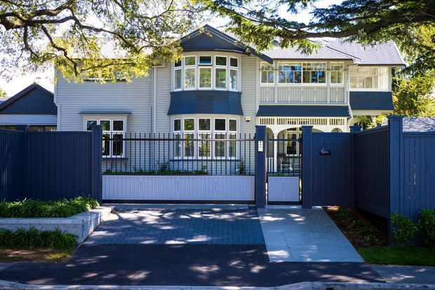 Street-facing exterior of a refined multi-storey residence with a gabled roof, pale horizontal weatherboard cladding, dark blue architectural accents, a projecting bay window, enclosed upper-level veranda, and coordinated dark fencing and pedestrian gates. A paved driveway and front garden frame the facade.