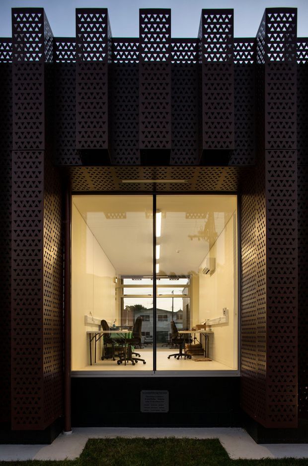 Exterior view of a compact contemporary building facade with a strongly patterned perforated screen in dark metal or masonry. Repeated projecting vertical fins create a fortress-like rhythm around a fully glazed entrance or office front. Warm interior lighting reveals a small workspace with desks and office chairs behind the glazing. The facade has geometric triangular perforations and a sheltered canopy above the entry.