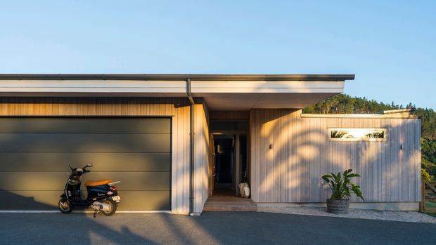 Front exterior view of a compact contemporary dwelling or ancillary building, featuring a flat low-pitched roof, attached single-car garage, recessed entry, and vertically clad timber-toned facade. A narrow horizontal window and dense vegetation are visible to the right, with a scooter parked in the driveway.