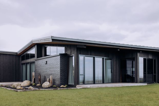 Exterior view of a low-slung contemporary house with dark vertical timber cladding, a shallow pitched metal roof, extensive floor-to-ceiling glazing, and a projecting dark brick volume. The home sits beside a broad lawn with minimalist rock landscaping under an overcast sky.