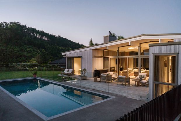 Contemporary single-storey pavilion-style home set against a wooded hillside, photographed at dusk. The building features a low-pitched flat roof, extensive floor-to-ceiling glazing, vertical grey timber cladding, and a large open-plan living area opening onto a timber pool deck. A rectangular swimming pool with glass balustrade dominates the foreground, while warm interior lighting creates an inviting connection between indoor and outdoor spaces.