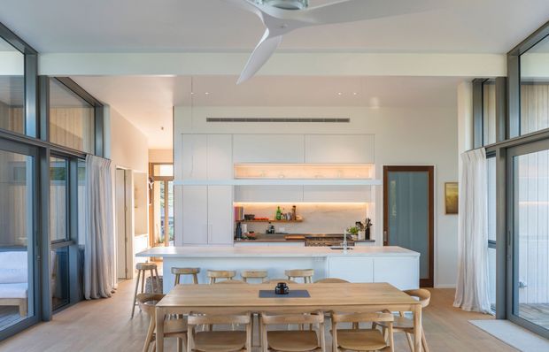 Bright open-plan kitchen and dining area with a large timber dining table in the foreground, a pale kitchen island with integrated seating, extensive floor-to-ceiling glazing, sheer curtains, white built-in cabinetry, and a contemporary ceiling fan. The space has a clean, warm, light-filled coastal feel.