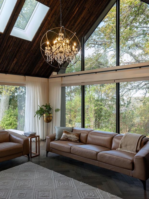 A warm, sophisticated living room with expansive floor-to-ceiling windows overlooking mature trees. The room features a large brown leather sectional sofa, patterned area rug, sheer curtains, a tall potted plant, and an ornate crystal chandelier suspended beneath a dramatic dark timber vaulted ceiling with skylights. Dark herringbone flooring and generous glazing create a strong connection to the surrounding landscape.