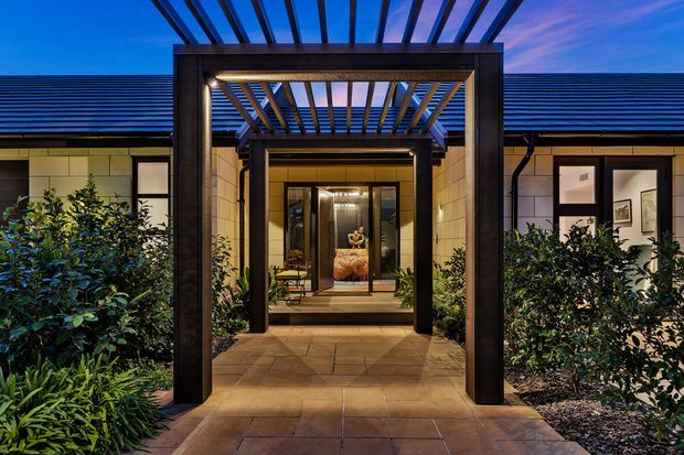 Dusk exterior view of a single-storey residence entry, framed by a dark timber pergola and deep portal structure. A paved path leads through landscaped planting to glazed double entry doors, with warm interior lighting visible beyond. The home has a low-pitched dark roof, pale masonry or rendered wall sections, and symmetrical windows.