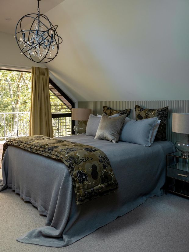 A refined bedroom with a blue-grey upholstered bed dressed in layered blue-grey bedding, patterned botanical cushions and a matching throw. The room has a dramatic sloped ceiling, a triangular black-framed window opening to a balcony, full-height neutral curtains, vertical ribbed wall panelling behind the bed, glass bedside tables, and an ornate spherical crystal chandelier. The palette is cool and muted with decorative, luxurious detailing.