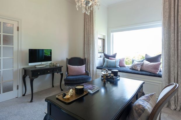 Elegant, traditionally styled living room or sitting room with a large dark wood table, upholstered dining chairs, a black accent armchair, and a television on a slender console. A bright bay-style window seat with cushions occupies the far wall, framed by full-length patterned curtains. The room has pale walls, carpeted flooring, a glazed-panel door, and a decorative crystal chandelier.