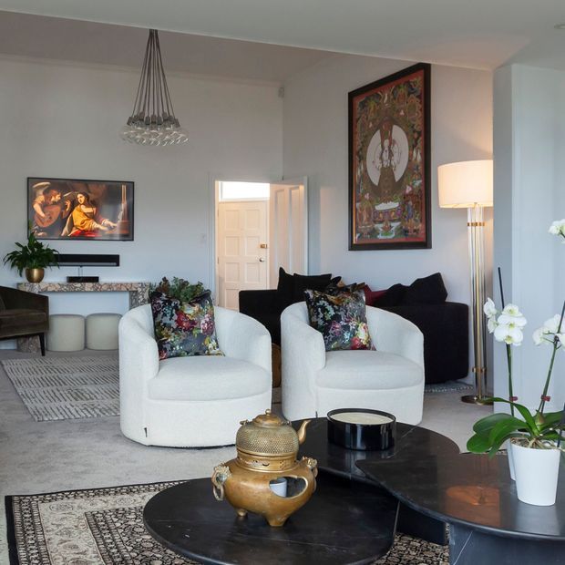 A refined contemporary living room with two curved white upholstered armchairs, a dark sofa, floral accent cushions, layered rugs, and a pair of black rounded coffee tables. The room features pale walls, a sculptural hanging chandelier, framed artwork, a floor lamp, built-in media cabinetry, and indoor orchids. A doorway opens to the hall beyond.