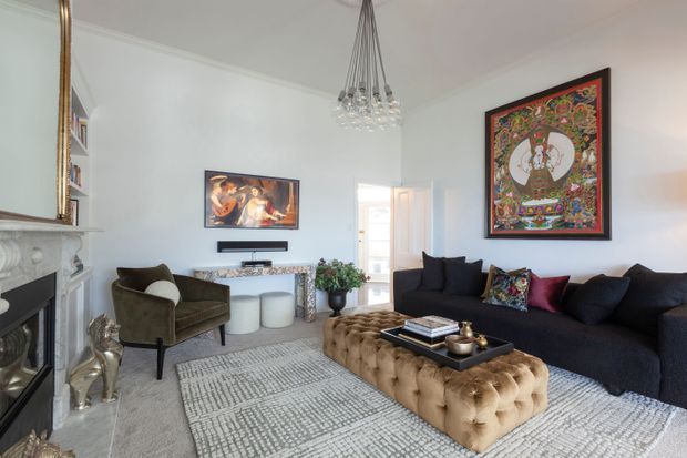 Elegant, light-filled living room with a dark sectional sofa, tufted tan ottoman, patterned area rug, marble fireplace, ornate wall mirror, framed artwork, a compact television, and a crystal pendant chandelier. The room combines classic architectural details with refined contemporary furnishings.
