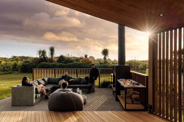 A covered outdoor entertaining area with a timber-lined ceiling, timber decking, vertical timber screening, and a dark-framed balustrade. The space contains a large outdoor sectional lounge, beanbag seating, a built-in outdoor fireplace or heater, and a serving console, opening onto a landscaped lawn and garden with sunset views.