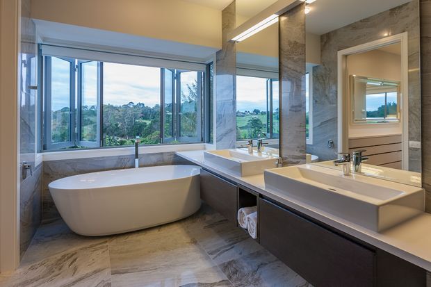 A spacious contemporary bathroom with a freestanding oval bathtub positioned beneath large corner windows, a double vanity with dark fixed cabinetry and integrated rectangular basins, and extensive grey-beige stone-look wall and floor finishes. Large mirrors reflect the vanity and doorway, while the windows provide views of leafy surroundings. The room is bright, refined, and luxurious.